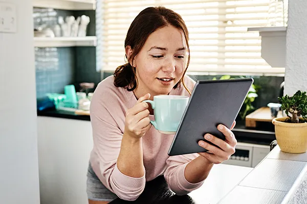 Woman holding a coffee cup and reading a tablet in her kitchen